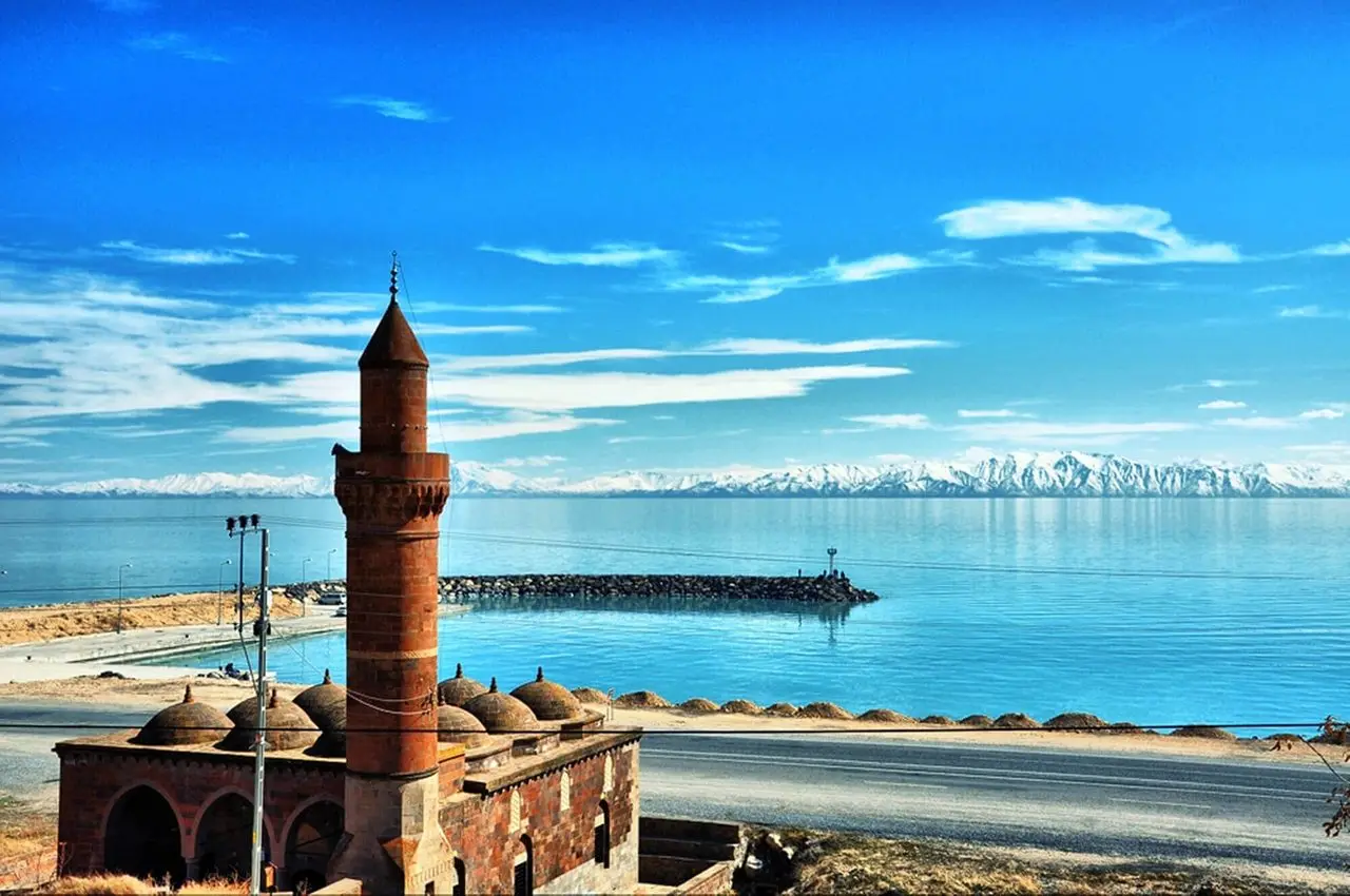 Adilcevaz’da Van Gölü kıyısı; ön planda taş caminin minaresi ve kubbeleri, arkada göl ve karlı dağlar